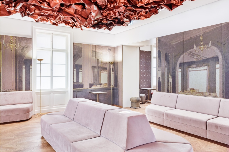 Auf dem Foto sehen wir den Tea Room im Museum der Heidi Horten Collection in Wien.
An der Decke hängt ein großes, rotes Kunstwerk von Hans Kupelwieser.
Es sieht aus wie zerknittertes Papier oder glänzender Stoff,
wurde aber aus Aluminium gemacht.
An der hinteren Wand gibt es viele runde Fenster – sie sehen aus wie Bullaugen.
Dahinter befinden sind kleine, kostbare Objekte aus der Sammlung von Heidi Horten.
Diese Vitrinenwand wurde von Markus Schinwald entworfen.
Auch die rosa Sitzmöbel in der Mitte des Raums gehören zu seinem Konzept.
Sie laden ein, sich hinzusetzen, zu schauen und zu entspannen.
Die Wände sind mit großen Stoffbildern bedeckt,
die an ein altes Café oder Schlosszimmer erinnern.