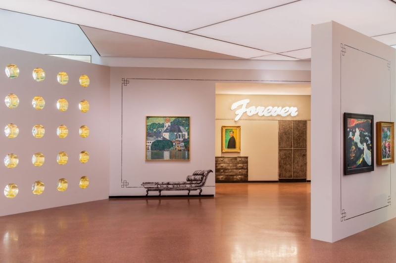 Auf dem Foto sieht man einen Teil der Ausstellung "Klimt⇄Warhol" im Museum der Heidi Horten Collection in Wien.
Links sieht man eine Wand mit runden Fenstern, die wie Bullaugen aussehen.
Diese Wand wurde von dem Künstler Markus Schinwald entworfen.
In der Mitte, direkt vor uns, hängt ein Gemälde von Gustav Klimt.
Darauf sieht man den Attersee, einen See in Österreich.
Das Bild zeigt Wasser, Häuser und Natur in hellen Farben.
Die Verzierungen an den Wänden und die gemalten Möbelstücke neben und unter den Kunstwerken hat ebenfalls Markus Schinwald gestaltet.
Hinten an der Wand, über einem Bild, leuchtet in großen weißen Buchstaben das Wort „Forever“.
Diese Leuchtschrift ist ein Kunstwerk von Tim Noble & Sue Webster.
Darunter hängt ein Selbstporträt von Edvard Munch.
Ganz rechts ragt eine weitere Wand schräg in den Raum hinein.
Dort hängen zwei Gemälde von Emil Nolde, die ebenfalls Teil der Sammlung sind.