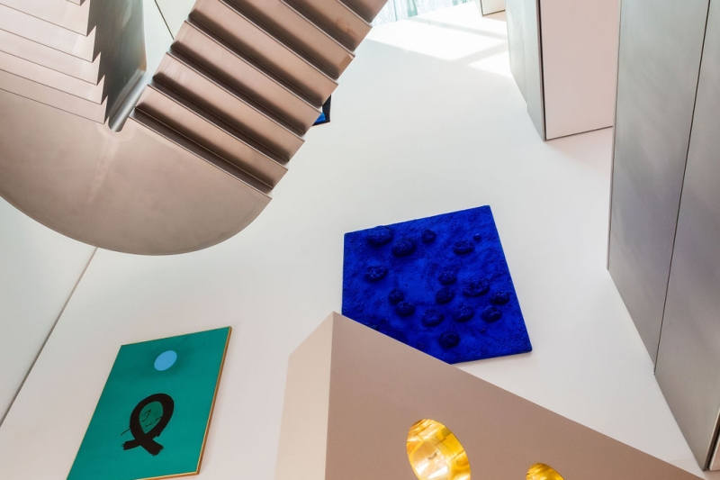 Auf dem Foto sieht man einen Blick nach oben im Museum der Heidi Horten Collection in Wien.
Man erkennt eine moderne Treppe aus Metall und große, helle Wände.
Links an der Wand hängt ein grünes Bild mit einem schwarzen Zeichen und einem blauen Kreis.
Dieses Kunstwerk heißt „Blue Orb“ und ist von Adolph Gottlieb.
Daneben, weiter oben, sieht man ein kräftig blaues Kunstwerk mit Schwämmen darauf.
Das ist das allererste Schwammrelief von Yves Klein.
Er hat echte Schwämme mit tiefblauer Farbe eingefärbt und auf die Leinwand geklebt.
Das Werk ist sehr besonders, weil Yves Klein für diese intensive blaue Farbe berühmt wurde.
Unten im Bild sieht man einen Teil der Bullaugenwand von Markus Schinwald.
Die runden, gold glänzenden Fenster gehören zu einem Gestaltungselement.