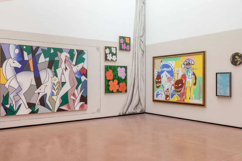 Auf dem Foto sieht man einen Teil der Ausstellung "Klimt⇄Warhol" im Museum der Heidi Horten Collection in Wien.
Die Aufnahme zeigt eine Ecke des Ausstellungsraums,
an der zwei Wände aufeinandertreffen. An diesen Wänden hängen mehrere bunte Gemälde.
Ganz links sieht man ein großes Bild von Roy Lichtenstein.
Es zeigt mehrere weiße Rehe in einer abstrakten Landschaft,
mit starken Farben und schwarzen Umrisslinien – typisch für Lichtensteins Stil, der an Comics erinnert.
Rechts daneben, übereinander gehängt, sieht man drei kleine Bilder mit bunten Blumen.
Diese Blumendrucke stammen von Andy Warhol. Sie sind sehr farbenfroh, mit einfachen Formen und starkem Kontrast.
In der rechten Ecke hängt ein großes, gemeinsames Gemälde von Jean-Michel Basquiat und Andy Warhol.
Das Bild ist wild, bunt und voller Energie.
Man erkennt Figuren, Schrift und Gesichter.
Rechts daneben sieht man einen weiteren Druck von Andy Warhol –
ein kleines, hellblaues Bild, auf dem man Elisabeth Taylor als Kleopatra sieht.
Darüber, rund in gold ist ein weiteres Werk von Warhol:
ein Porträt von Jackie Kennedy, der früheren First Lady der USA.
Ein aufgemalter, grauer Vorhang trennt die beiden Wände optisch und erinnert an ein Theater oder eine Bühne.
Auch dieser Teil der Wandgestaltung gehört zum inszenierten Ausstellungsdesign von Markus Schinwald.