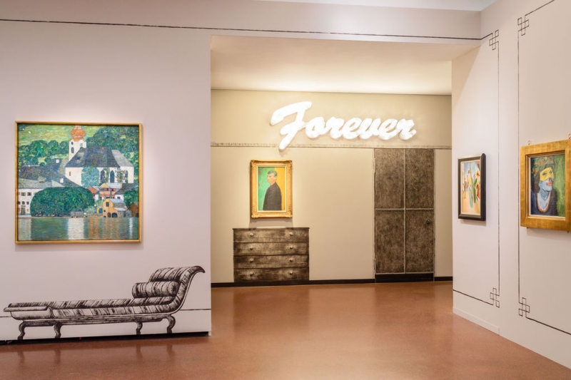 Auf dem Foto sieht man einen Teil der Ausstellung "Klimt⇄Warhol" im Museum der Heidi Horten Collection in Wien.
Links, hängt ein Gemälde von Gustav Klimt.
Darauf sieht man den Attersee, einen See in Österreich.
Das Bild zeigt Wasser, Häuser und Natur in hellen Farben.
Die Verzierungen an den Wänden und die gemalten Möbelstücke neben und unter den Kunstwerken hat ebenfalls Markus Schinwald gestaltet.
Hinten an der Wand, über einem Bild, leuchtet in großen weißen Buchstaben das Wort „Forever“.
Diese Leuchtschrift ist ein Kunstwerk von Tim Noble & Sue Webster.
Darunter hängt ein Selbstporträt von Edvard Munch.