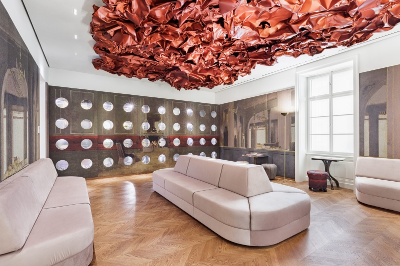 Auf dem Foto sehen wir den Tea Room im Museum der Heidi Horten Collection in Wien.
An der Decke hängt ein großes, rotes Kunstwerk von Hans Kupelwieser.
Es sieht aus wie zerknittertes Papier oder glänzender Stoff,
wurde aber aus Aluminium gemacht.
An der hinteren Wand gibt es viele runde Fenster – sie sehen aus wie Bullaugen.
Dahinter befinden sind kleine, kostbare Objekte aus der Sammlung von Heidi Horten.
Diese Vitrinenwand wurde von Markus Schinwald entworfen.
Auch die rosa Sitzmöbel in der Mitte des Raums gehören zu seinem Konzept.
Sie laden ein, sich hinzusetzen, zu schauen und zu entspannen.
Die Wände sind mit großen Stoffbildern bedeckt,
die an ein altes Café oder Schlosszimmer erinnern.