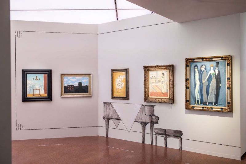 Auf dem Foto sieht man einen Teil der Ausstellung "Klimt⇄Warhol" im Museum der Heidi Horten Collection in Wien.
Man sieht eine Wand mit mehreren Gemälden. Links sind zwei surrealistische Bilder von René Magritte.
Unter den Gemälde mittig befindet sich eine Verzierung in Form eines Flügels. Die Verzierungen an den Wänden und die gemalten Möbelstücke hat der Künstler Markus Schinwald gestaltet.